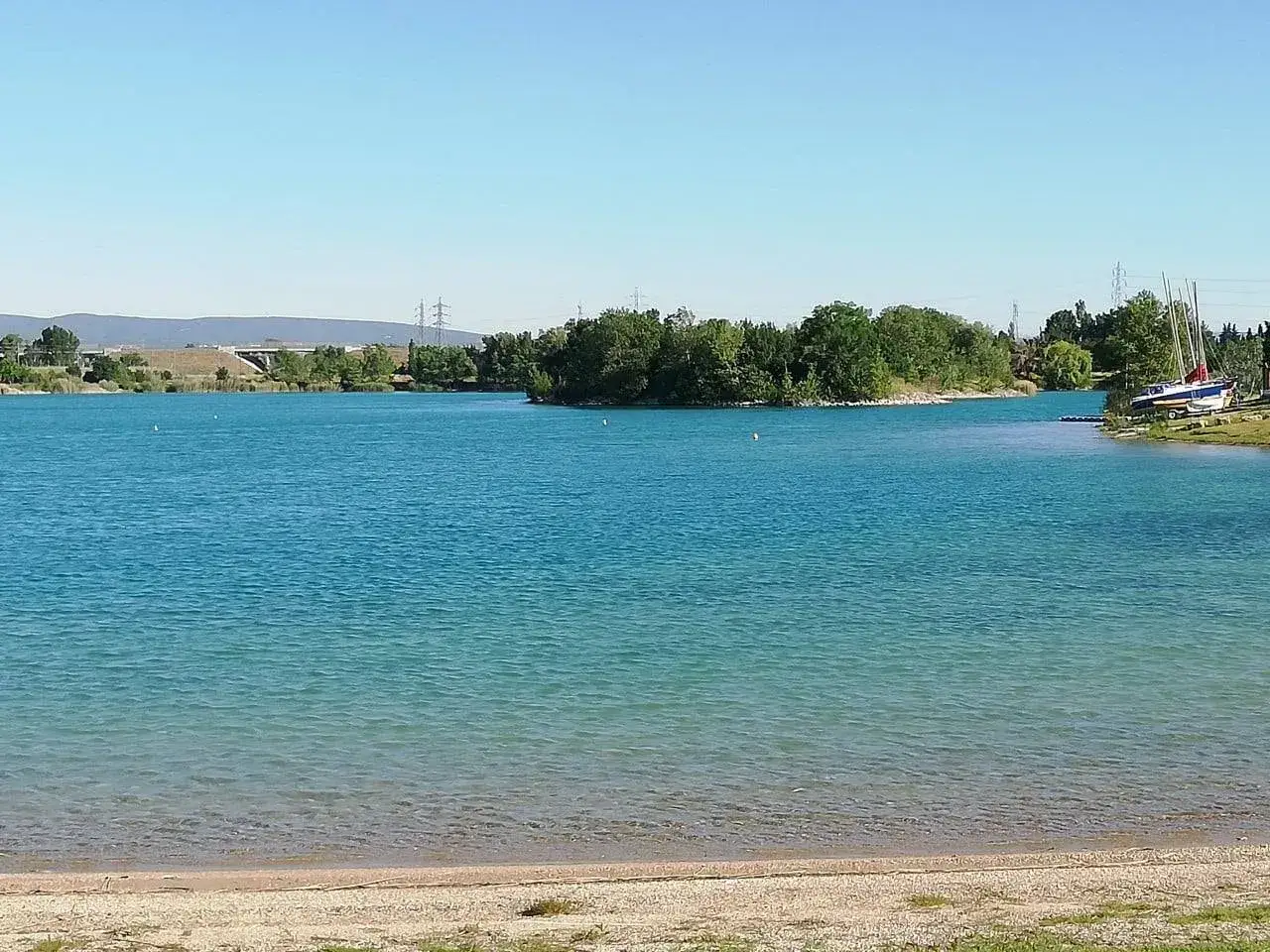 Vue paisible du lac de Lapalud, avec des voiliers amarr&eacute;s sur la rive et une &icirc;le verdoyante au loin sous un ciel bleu clair.