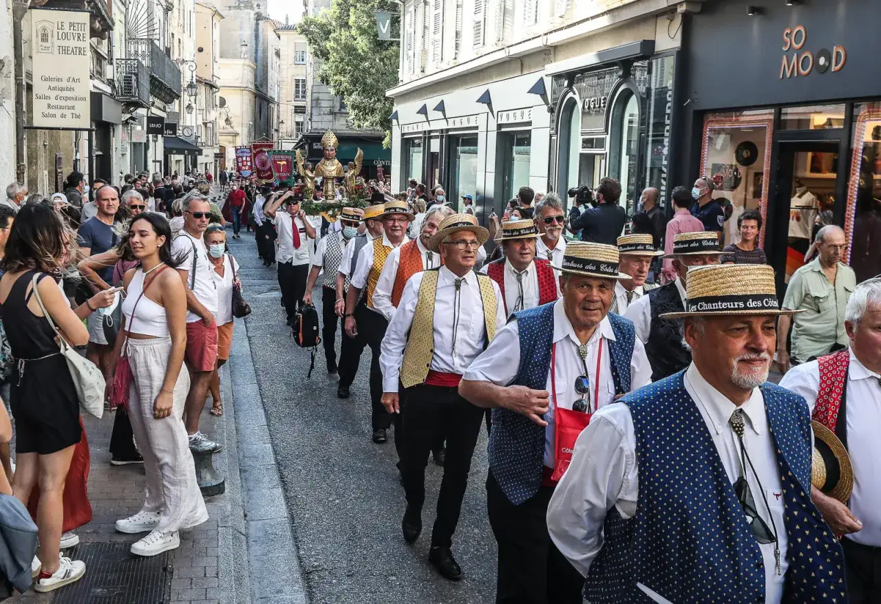 C&eacute;l&eacute;bration du ban des vendanges &agrave; Avignon. Un groupe d'hommes en tenues traditionnelles, chapeaux de paille et gilets color&eacute;s d&eacute;file dans la rue, portant une statue dor&eacute;e.