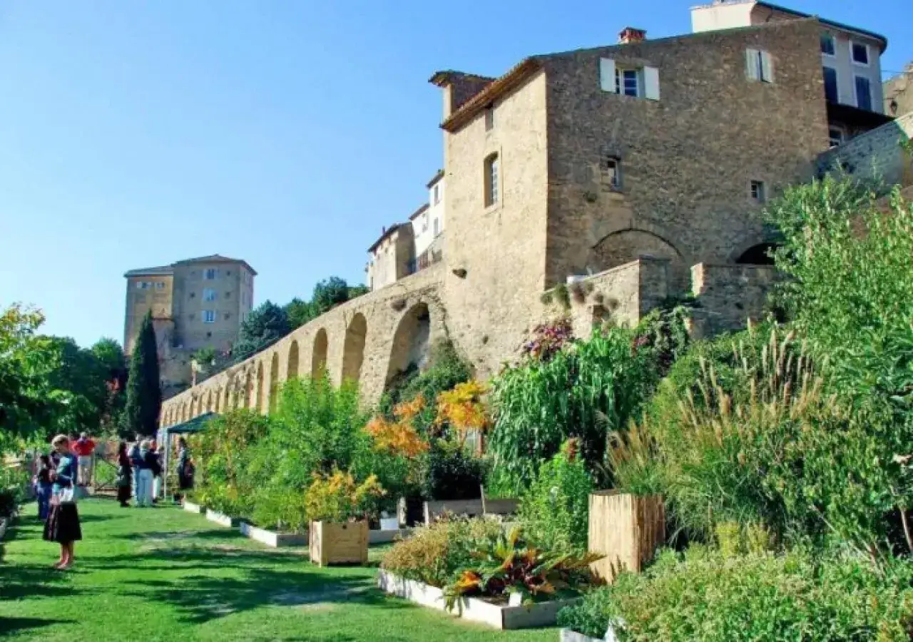 Jardin fleuri devant le ch&acirc;teau de Lauris, avec ses remparts vo&ucirc;t&eacute;s et ses b&acirc;timents en pierre sous un ciel bleu.