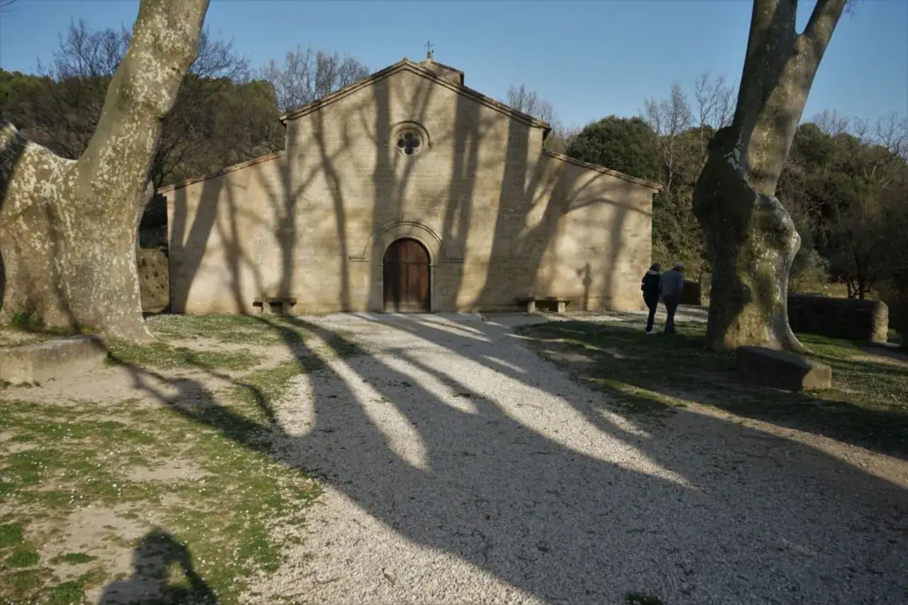 L'&eacute;glise de Vaugines, baign&eacute;e de soleil, projette de longues ombres d'arbres sur son parvis. Deux visiteurs admirent sa fa&ccedil;ade romane.
