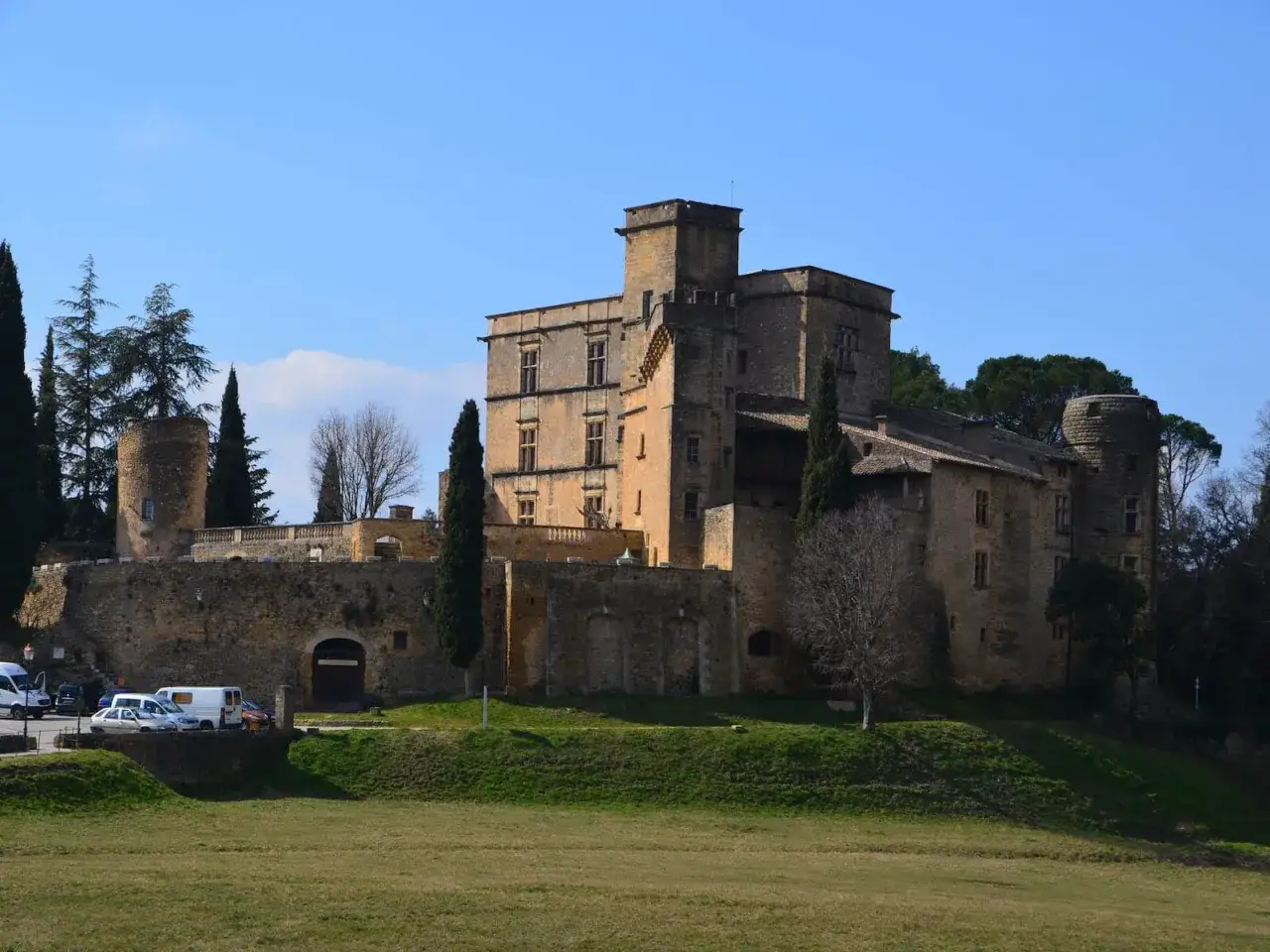 Le majestueux Ch&acirc;teau de Lourmarin se dresse sur une colline, entour&eacute; de cypr&egrave;s et d'arbres. Un ciel bleu clair domine le paysage.