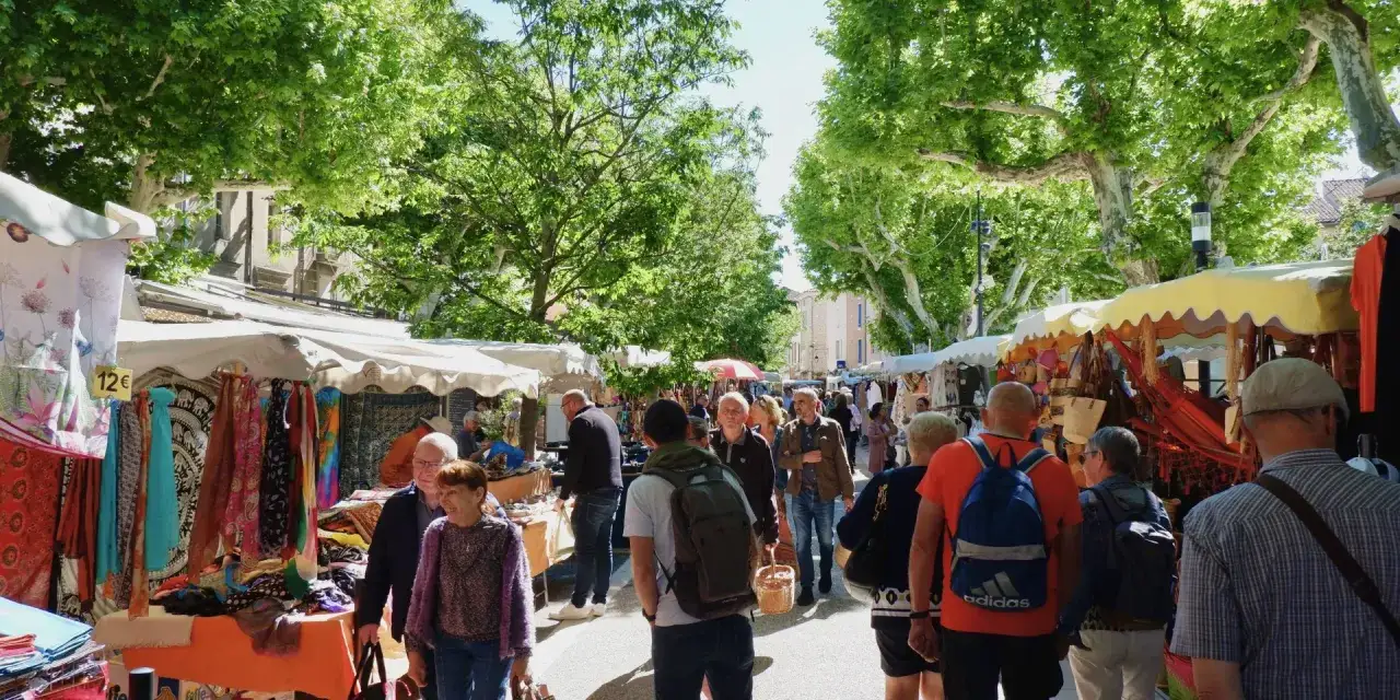 Ambiance anim&eacute;e au march&eacute; de Vaison-la-Romaine, sous les platanes, avec des &eacute;tals color&eacute;s de tissus et d'artisanat.