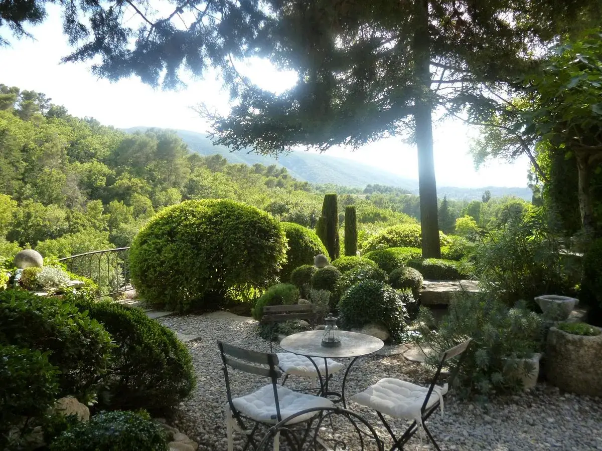 Le jardin de la louve, un havre de paix verdoyant avec des arbustes taill&eacute;s, une table et des chaises invitant &agrave; la d&eacute;tente, surplombant une vall&eacute;e luxuriante.