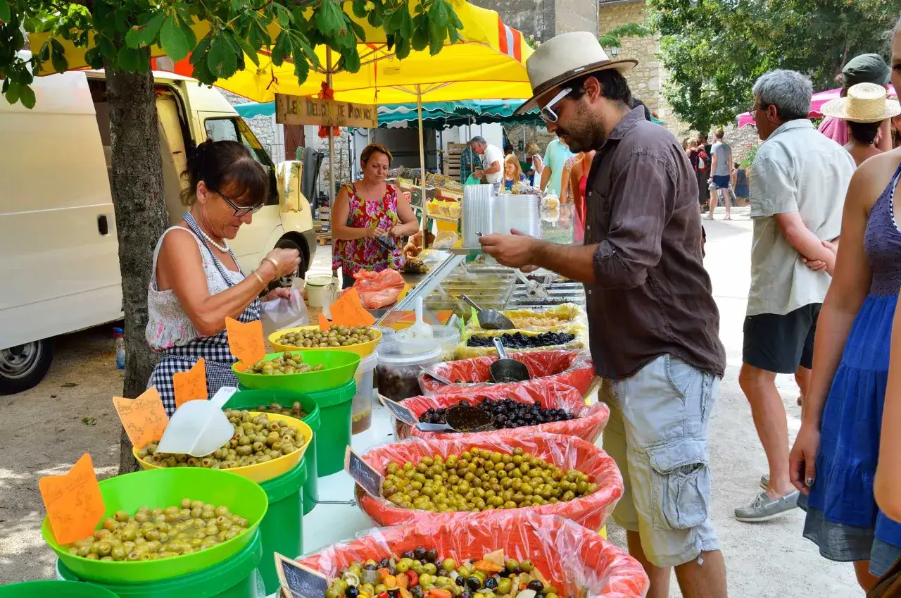 Un homme choisit des olives au march&eacute;, sous un soleil radieux. Les &eacute;tals d&eacute;bordent de fruits et l&eacute;gumes, offrant une belle palette de couleurs.