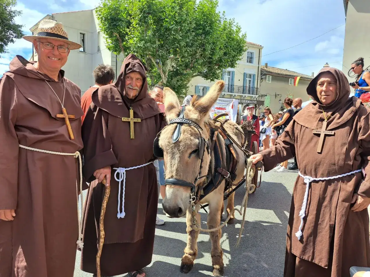 Trois moines et un âne participent à la fête de la veraison, une procession colorée dans un village ensoleillé.