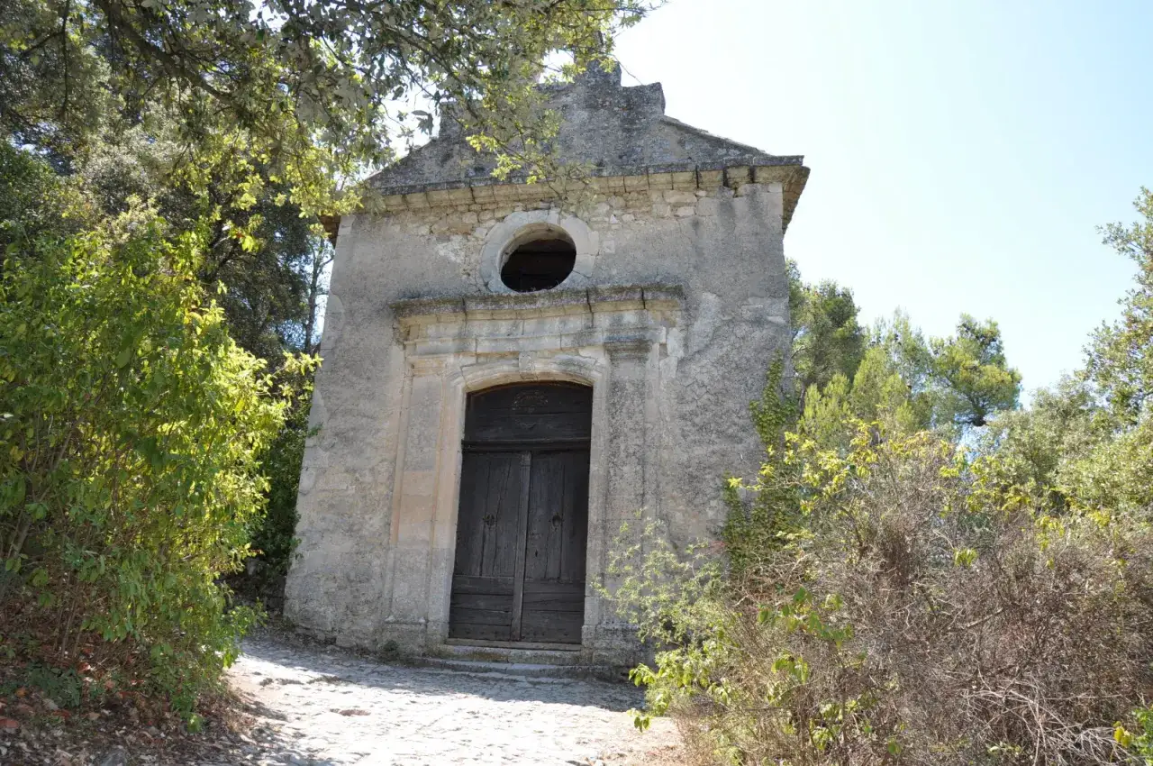 Chapelle Notre-Dame d'Alidon, petite b&acirc;tisse de pierre grise avec une porte en bois sombre, entour&eacute;e de verdure luxuriante sous un ciel bleu.