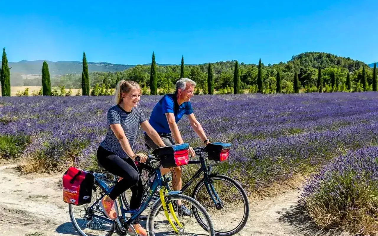 Couple souriant faisant du vélo à travers un champ de lavande en Provence, sous un ciel bleu éclatant.