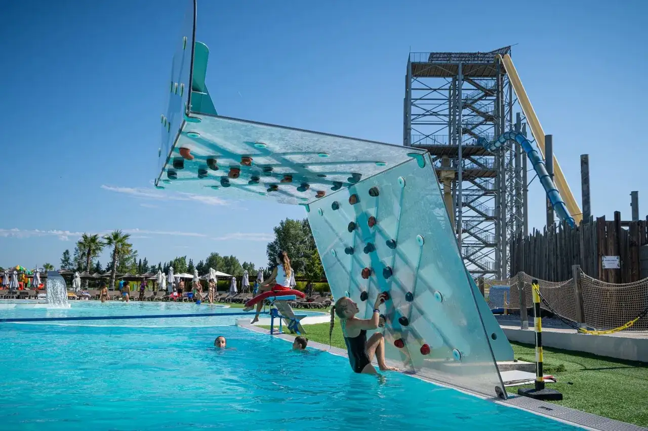 Une jeune fille escalade un mur transparent au bord d'une piscine, près d'une tour de toboggan aquatique, dans un parc aquatique qui ressemble à une île de loisirs.