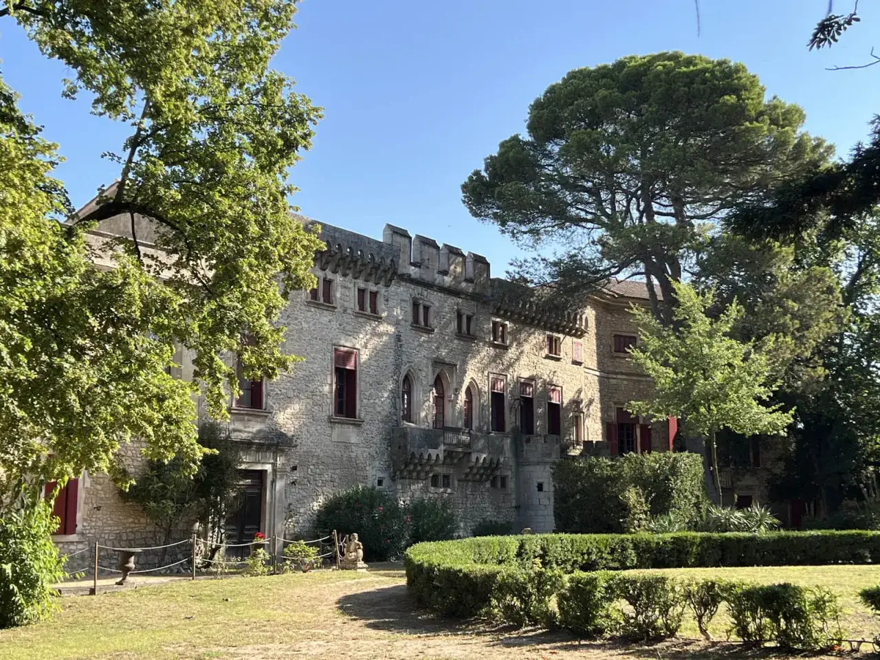 Le chateau de Thezan, une b&acirc;tisse de pierre aux volets rouges, se dresse fi&egrave;rement sous un ciel bleu, entour&eacute; d'arbres verdoyants et d'un jardin soign&eacute;.