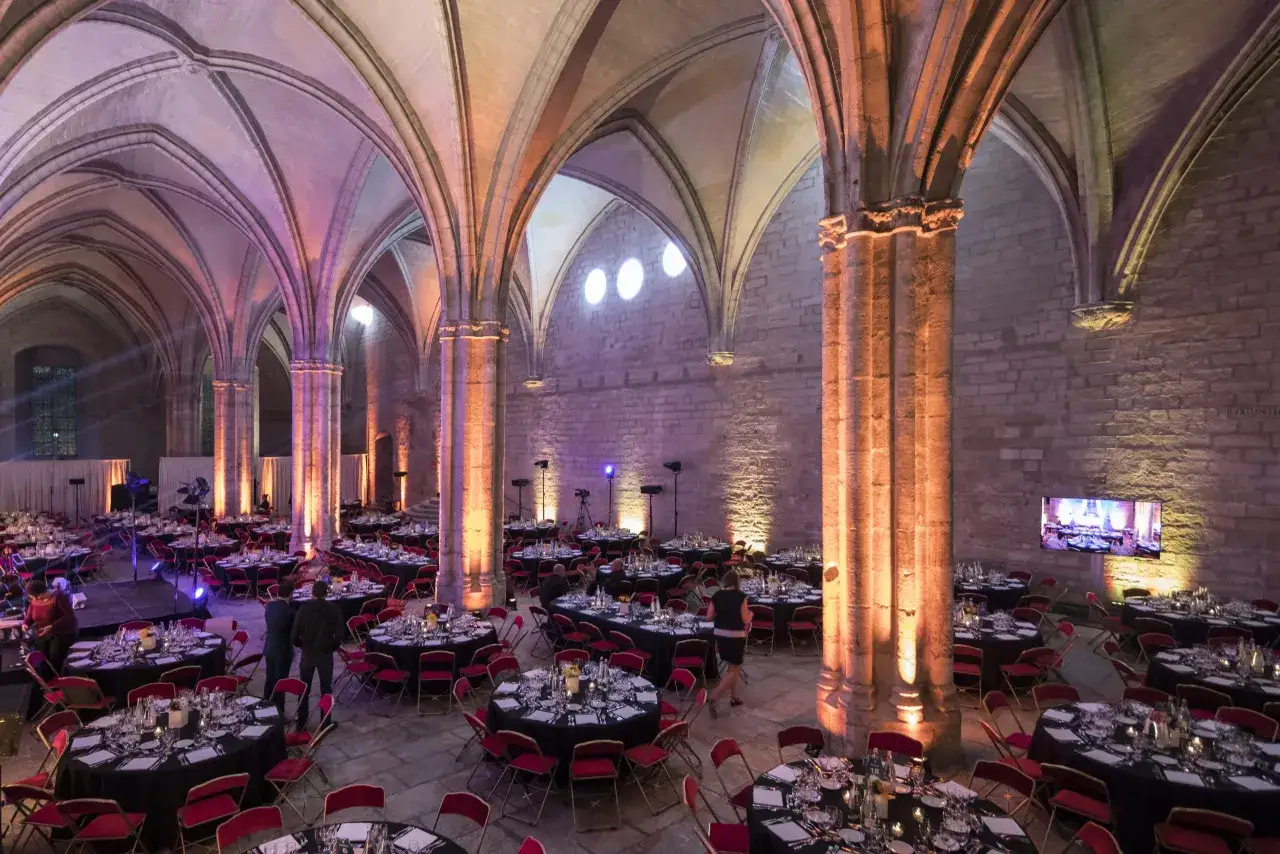 Int&eacute;rieur du Palais des Papes &agrave; Avignon, une salle vo&ucirc;t&eacute;e orn&eacute;e de lumi&egrave;res color&eacute;es, pr&ecirc;te pour un banquet avec des tables rondes dress&eacute;es.