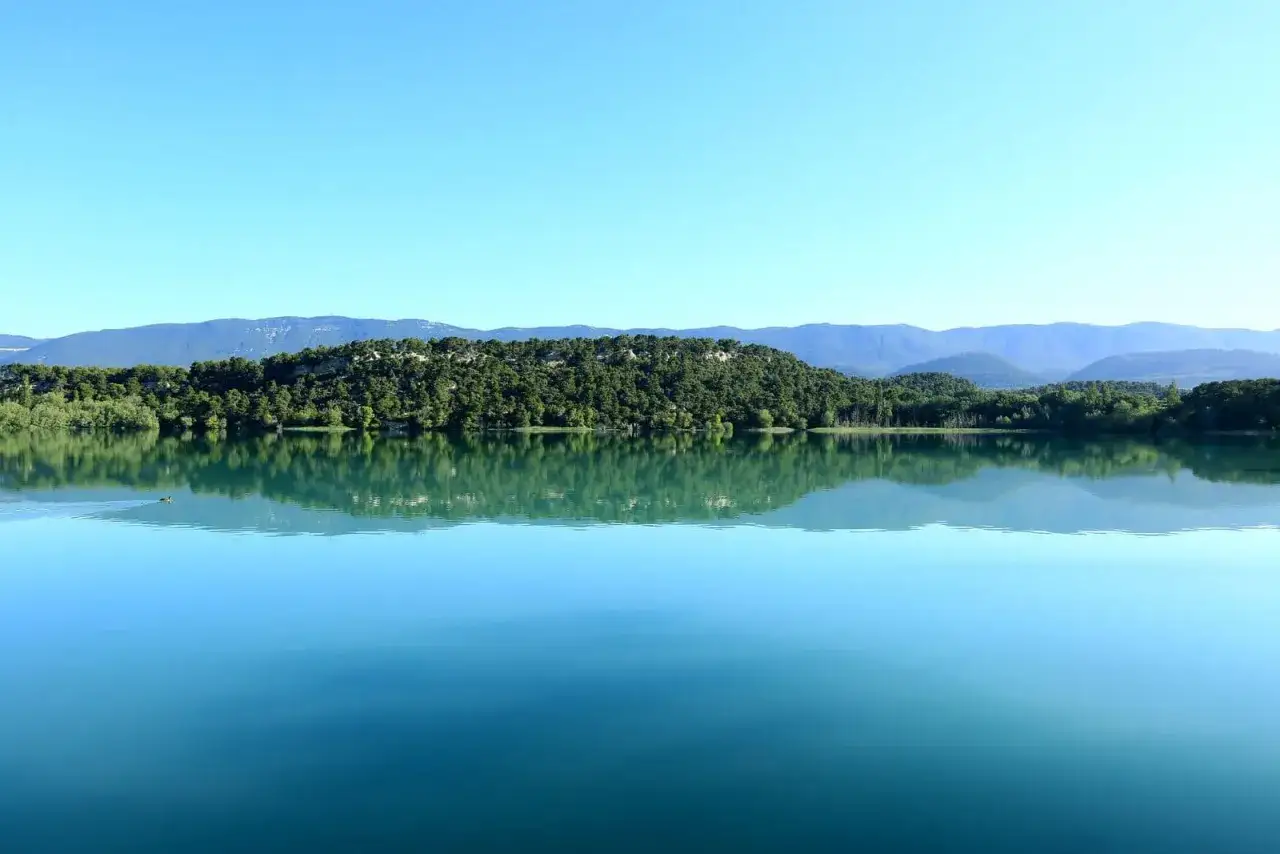Calme et serein, le lac de Bonnieux refl&egrave;te une colline verdoyante sous un ciel bleu azur. Une l&eacute;g&egrave;re ondulation sur l'eau r&eacute;v&egrave;le la pr&eacute;sence d'un canard.