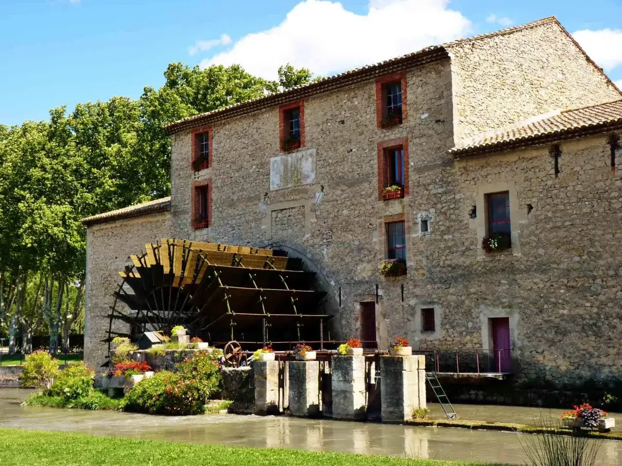 Vieux moulin en pierre avec une grande roue à aubes, entouré de verdure et de fleurs. Les taillades semblent figées dans le temps.