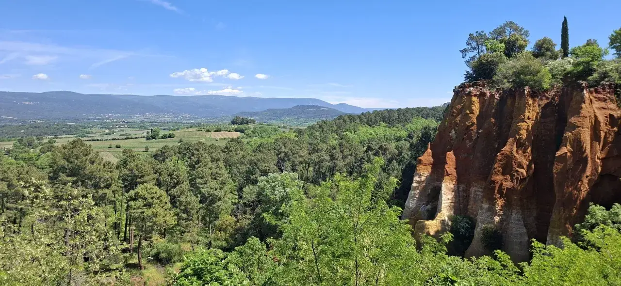 Paysage de Provence avec des falaises ocres spectaculaires, des for&ecirc;ts verdoyantes et des collines bleut&eacute;es au loin. Le sentier des ocres offre une vue imprenable.