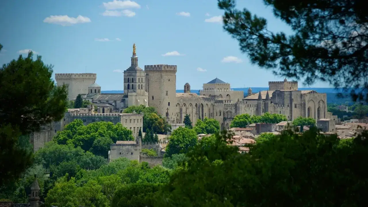 Vue panoramique de la cit&eacute; des papes, avec le Palais des Papes imposant, entour&eacute; de verdure luxuriante et sous un ciel bleu parsem&eacute; de nuages.