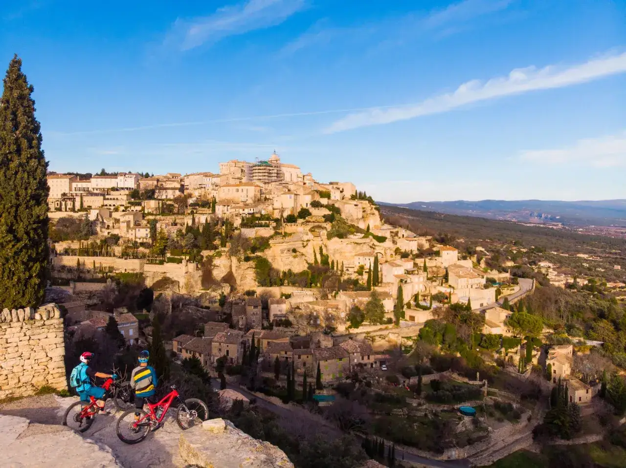 Deux cyclistes contemplent la ville de murs perchée sur une colline, sous un ciel bleu.