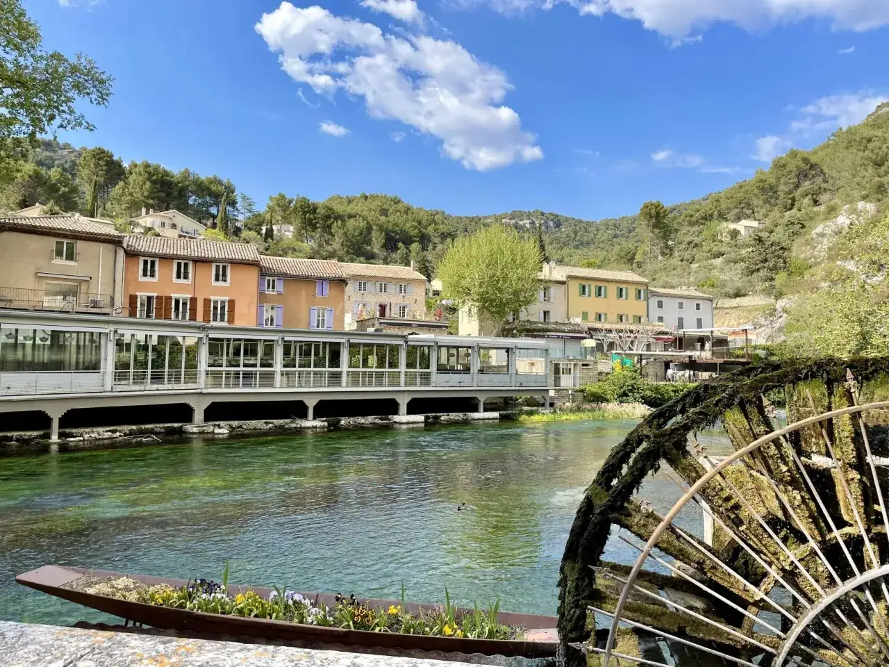 Le gouffre de Fontaine-de-Vaucluse vu depuis la rivi&egrave;re, avec une roue &agrave; aubes ancienne et des maisons color&eacute;es en arri&egrave;re-plan.