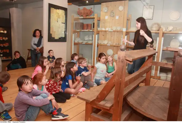 Visite d'un musée à Apt : des enfants écoutent attentivement une guide devant des étagères remplies d'objets anciens.