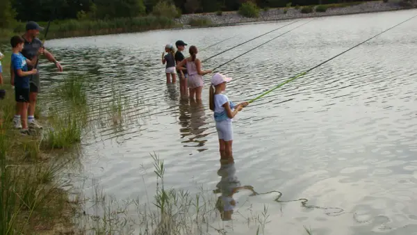 Des enfants pêchent dans le lac du Bartras, guidés par un adulte. L'eau reflète le ciel et la végétation.