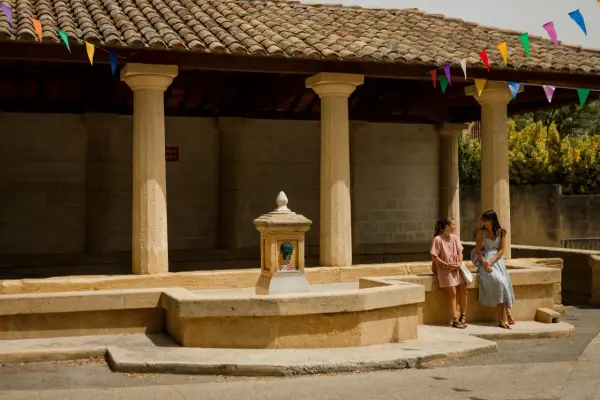 Deux femmes discutent près d'une fontaine sous un auvent orné de fanions colorés, ambiance de marché à Rochefort-du-Gard.