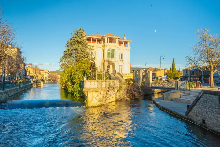 L'eau vive de la Sorgue traverse L'Isle-sur-la-Sorgue, bordée de maisons et d'un bâtiment blanc imposant.