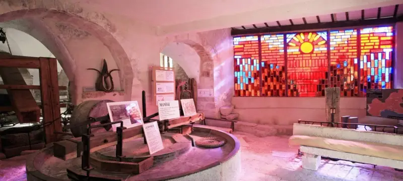 Dans le musée du vitrail Gordes, une roue de moulin ancienne côtoie un vitrail éclatant de soleil et de couleurs chaudes.